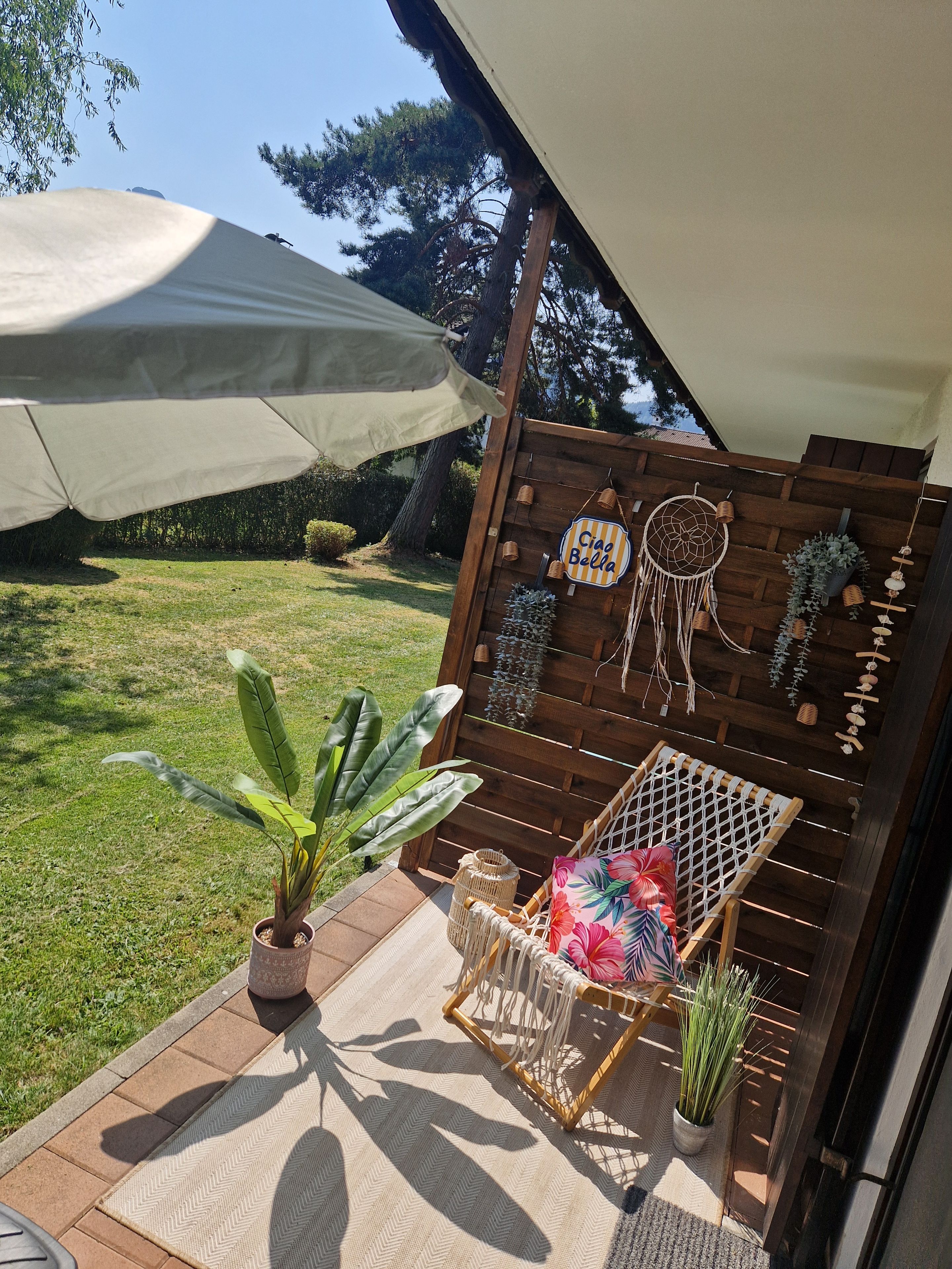 Una zona patio con recinzione in legno, una sedia in macramè, un decoro a forma di acquilone appeso e una pianta in vaso.