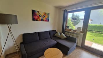 Ein modernes Wohnzimmer mit einer Eckcouch, einem runden Holztisch, einer Stehlampe und einem großen Fenster mit Blick auf Grünflächen.