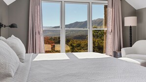 Une chambre avec un grand lit, une table de chevet, une lampe et une vue sur les montagnes grâce à de grandes fenêtres.
