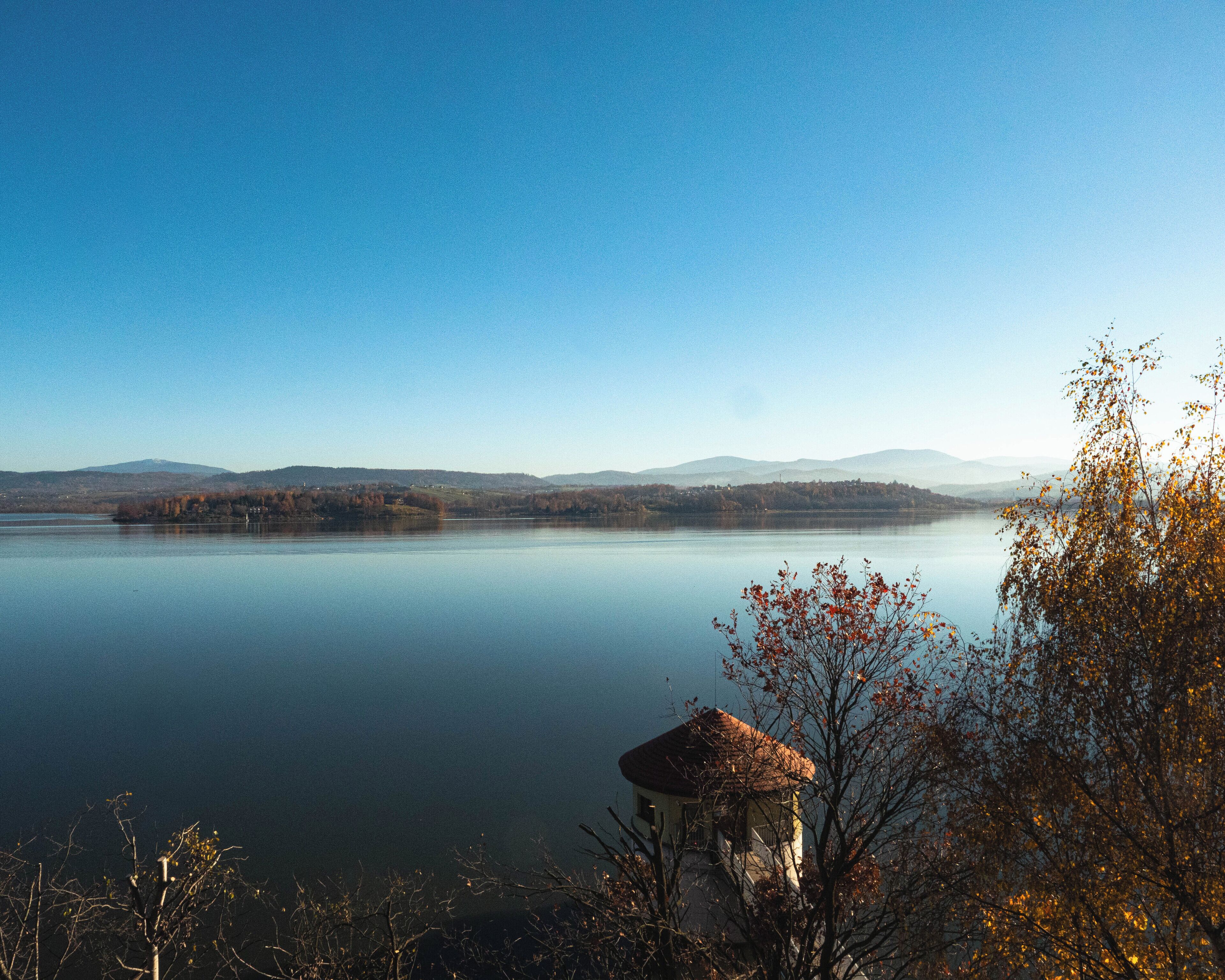 A tranquil lake with a clear sky, surrounded by hills and a small building with a thatched roof in the foreground.