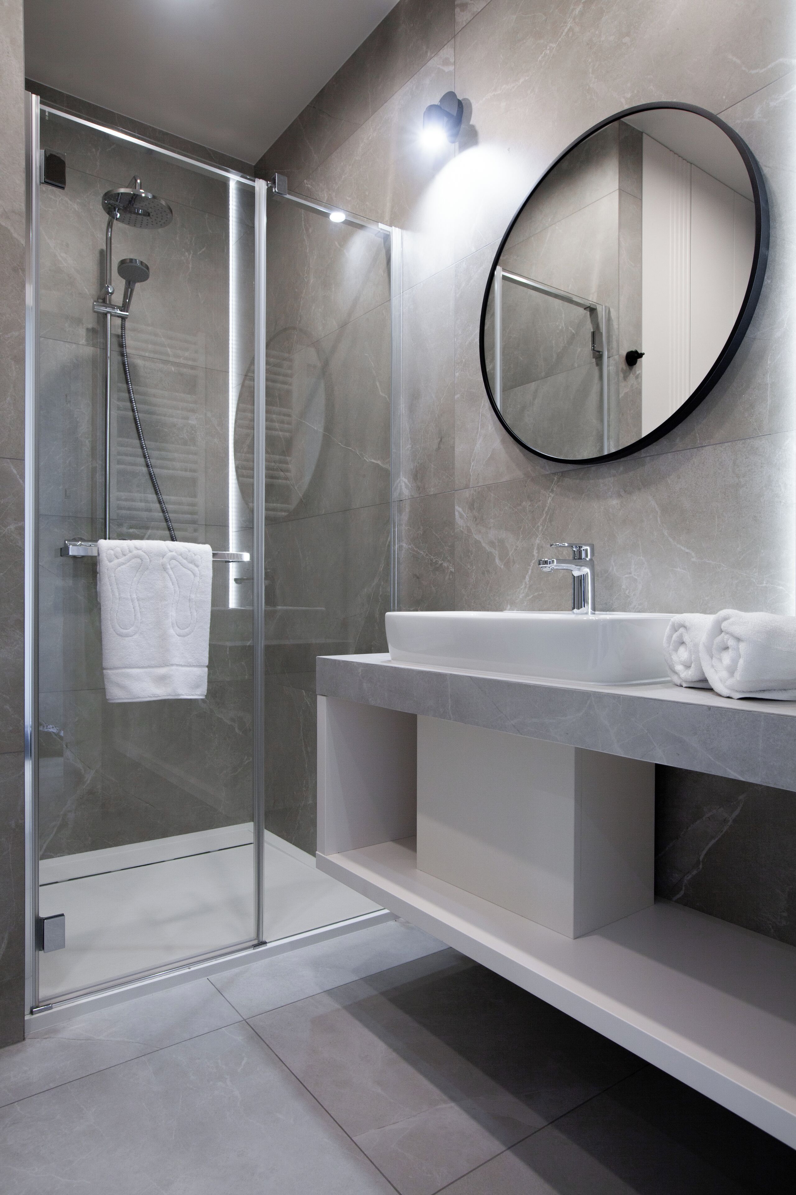 Een moderne badkamer met een douchecabine van glas, een ronde spiegel en een wit wastafelmeubel met een marmeren blad.