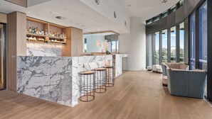 Een moderne barruimte met marmeren aanrechtbladen en krukken, een houten kast met flessen, en een groot raam met uitzicht over de stad.