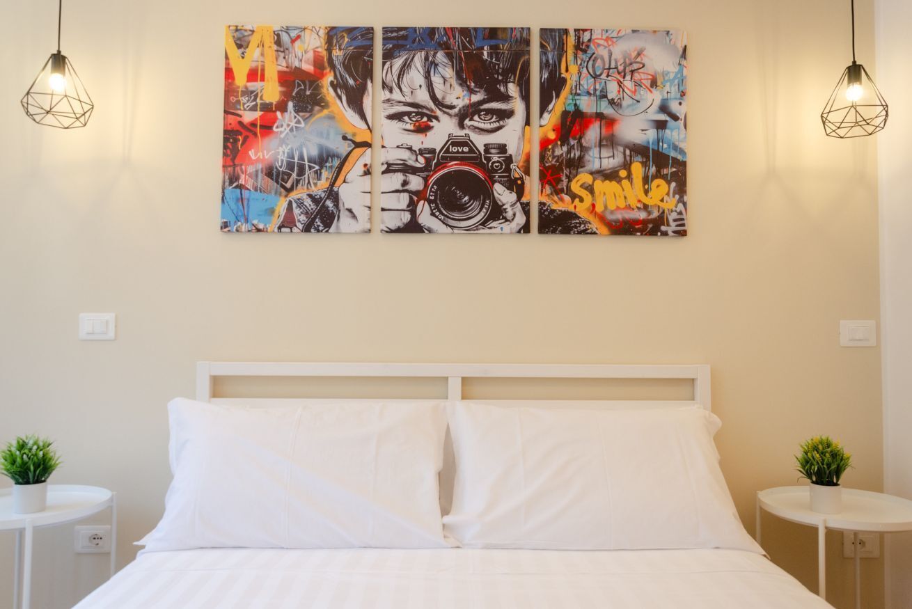 A hotel room with a white bed, two bedside tables with plants, and a large abstract painting on the wall.