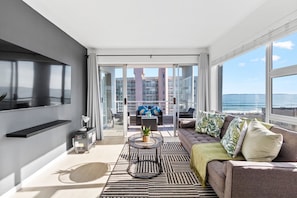 A modern living room with a large TV, a sofa, a coffee table, and a view of the ocean.