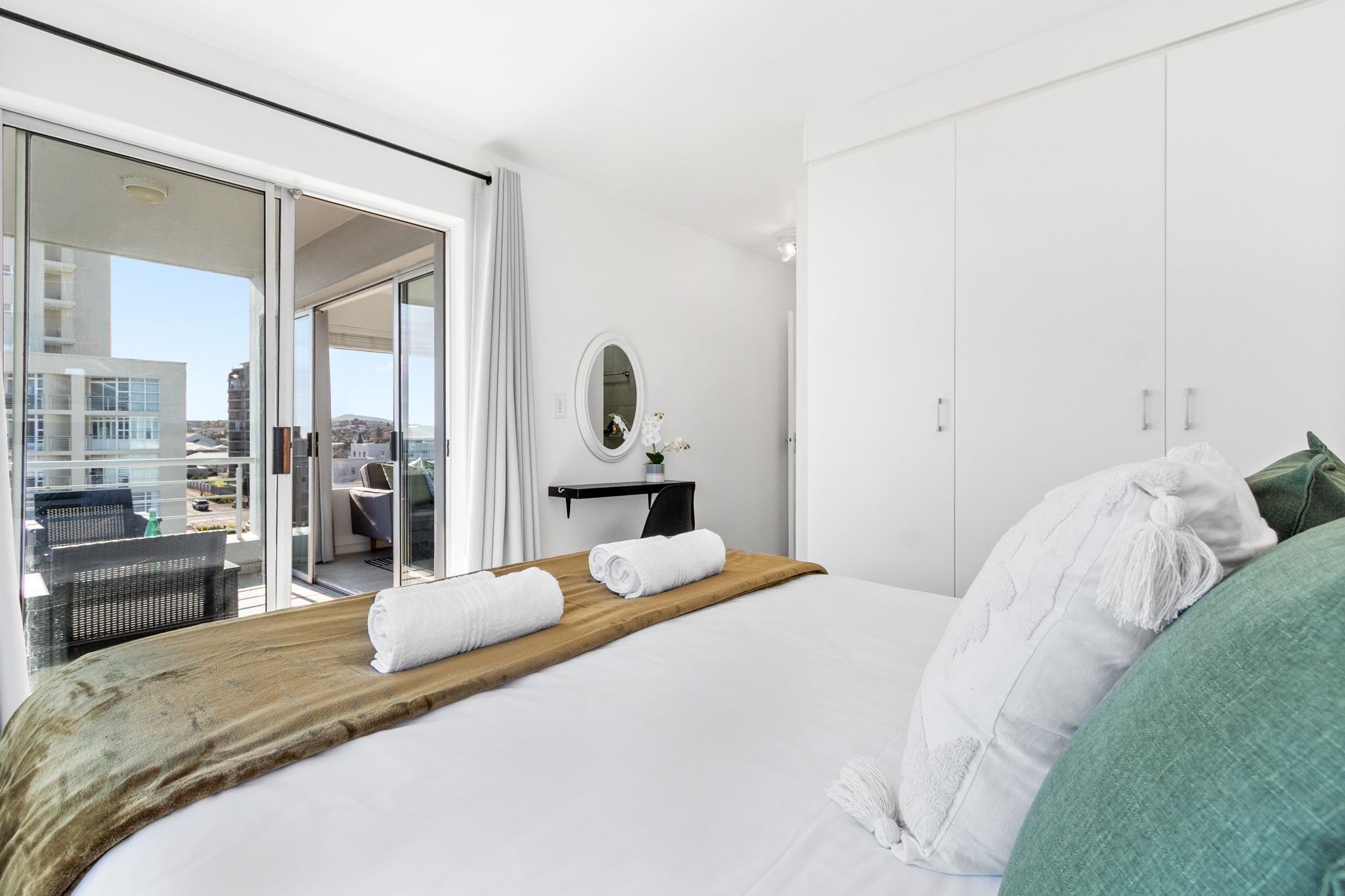 Une chambre d’hôtel moderne avec un grand lit, une tête de lit en bois, un petit bureau avec un miroir et une vue sur la ville à travers de grandes portes vitrées.