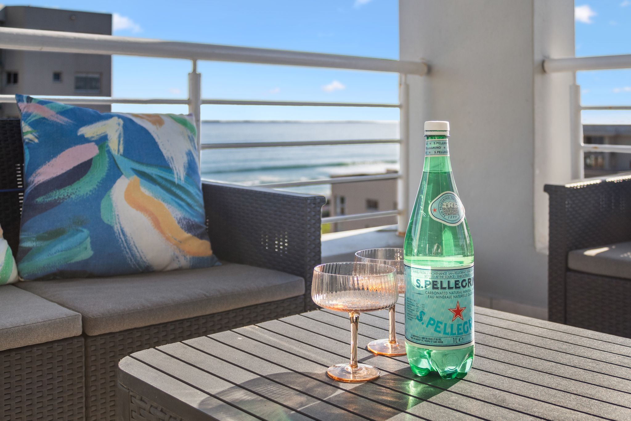 Una botella de agua Pellegrino y dos vasos sobre una mesa de madera con vista al océano.