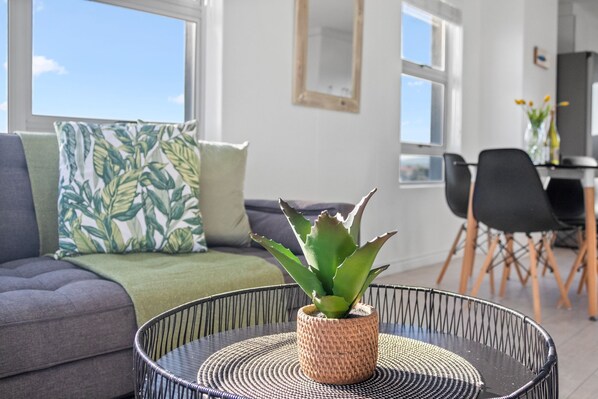 Un salon moderne avec un canapé gris, un coussin à motifs, une table ronde agrémentée d’un élément décoratif tissé, et une vue sur le ciel par de grandes fenêtres.