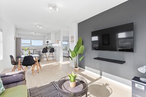 A modern living room with a dining area, a large potted plant, a flat-screen TV mounted on the wall, and a coffee machine on a table. - Ocean View B503 (Cape Town)
