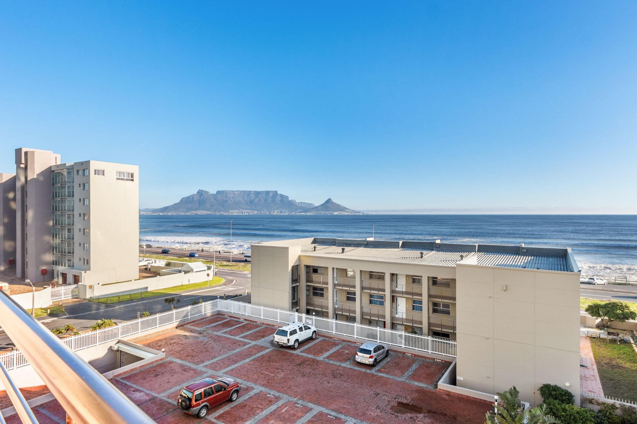A parking area with several cars, a building with a flat roof, and a view of the ocean and a distant mountain.
