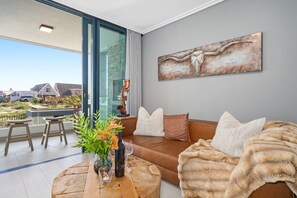 A modern living room with a large window, a brown leather sofa, a wooden coffee table, and a decorative metal artwork. - Azul Terraces 101 (Cape Town)