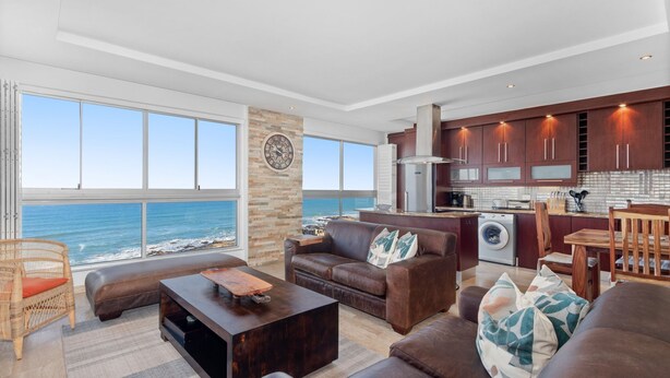 A spacious living area with a view of the ocean, a stone pillar, and a modern kitchen with wooden cabinets. - Silverwind - Blouberg Heights 1501 (Cape Town)