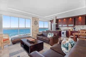 A modern living room with a large window overlooking the ocean, a kitchen with wooden cabinets, and a stone accent wall.