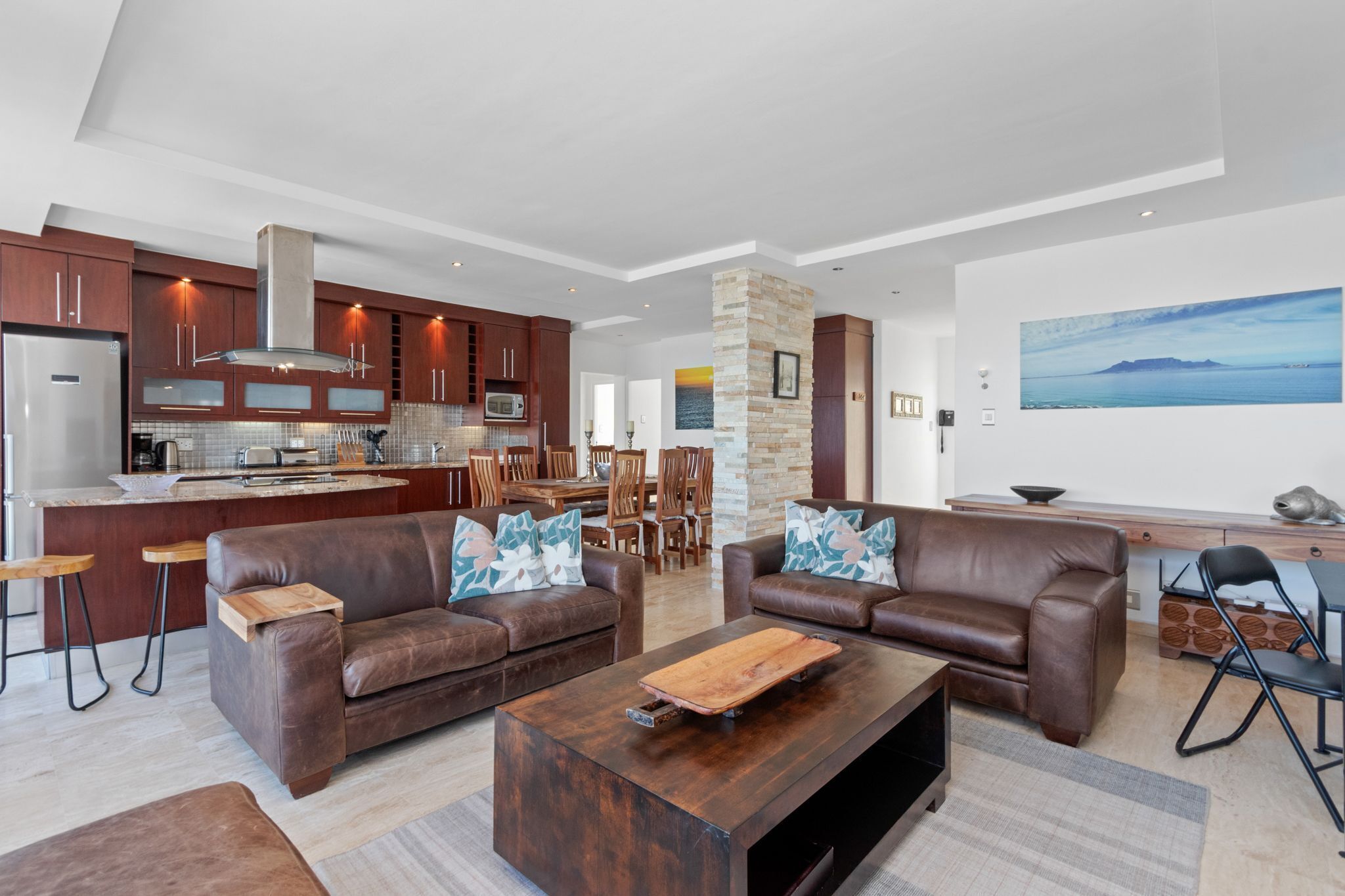 Une cuisine moderne avec des meubles en bois foncé, un réfrigérateur en acier inoxydable et un îlot central. Le salon comprend deux canapés en cuir marron ornés de coussins à motifs floraux, une table basse en bois et un coin repas avec des chaises en bois.