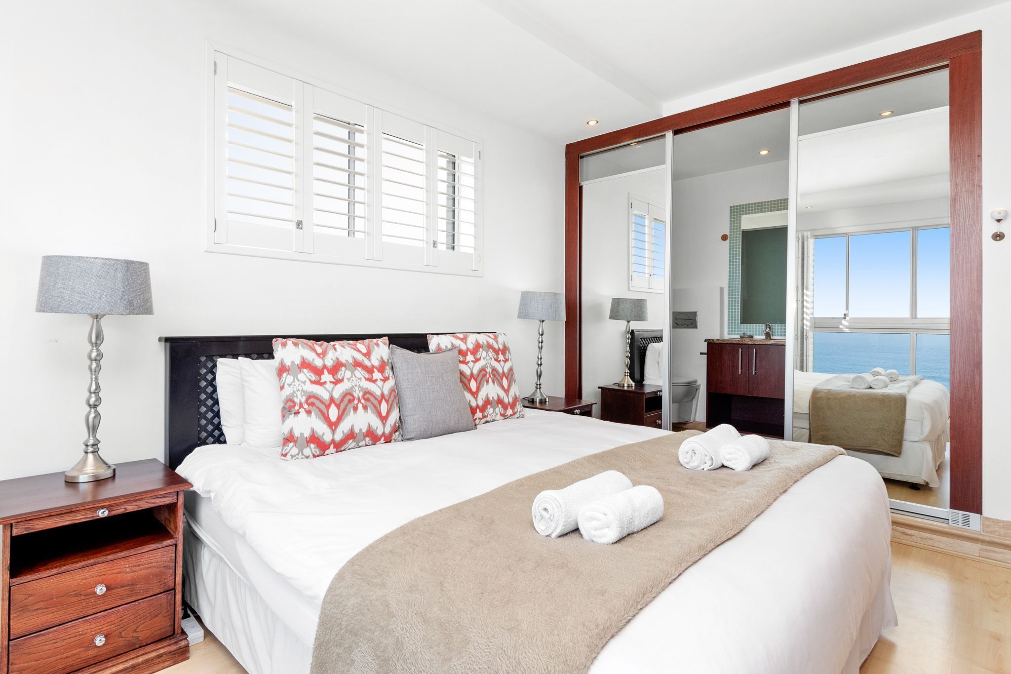 A hotel room with a bed, bedside lamps, a wooden nightstand, a large mirror, and a view of the sea through a sliding door.
