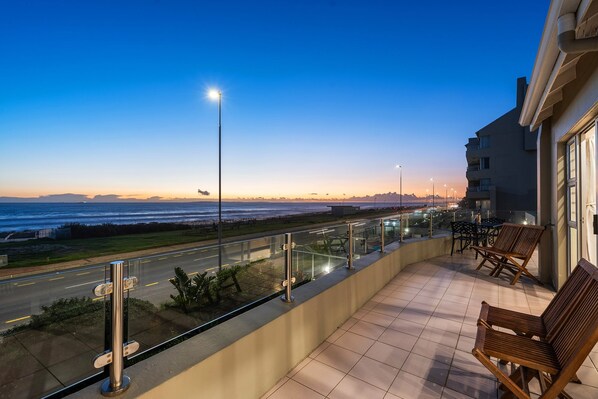 A balcony with a view of the ocean, a road, and a building with outdoor seating.