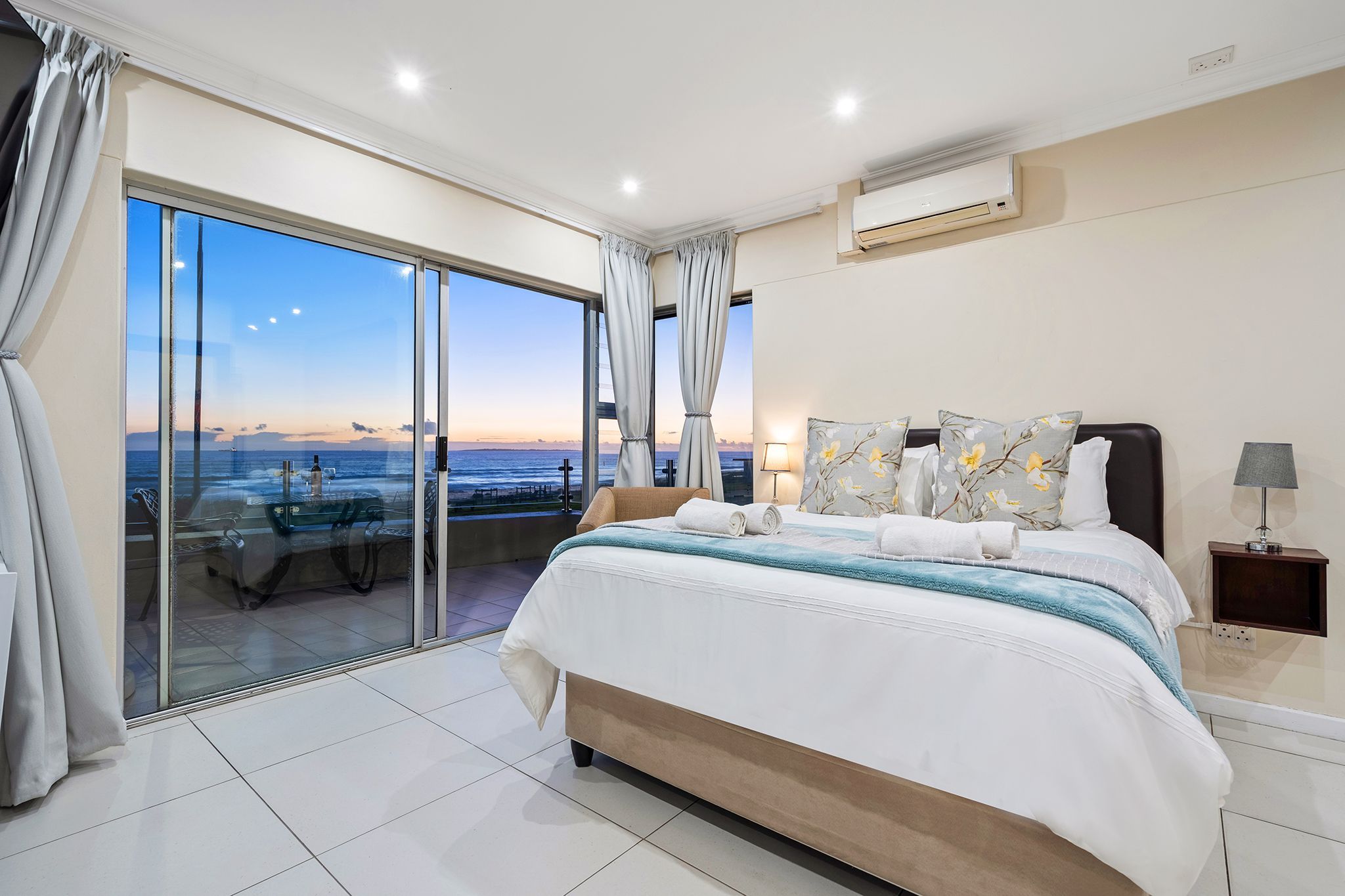 A bedroom with a large bed, a nightstand, a lamp, and a view of the ocean through a sliding glass door.