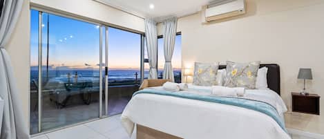 A bedroom with a large bed, a nightstand, a lamp, and a view of the ocean through a sliding glass door.