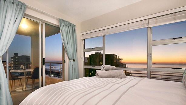 A hotel room with a large bed, a balcony with a table and chairs, and a view of the city at sunset. - Ocean View A603 (Cape Town)