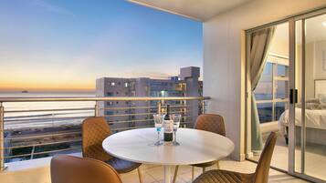 A balcony with a table and chairs, a bottle of wine, and a view of the sea and buildings.