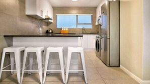 A modern kitchen with a central island, white bar stools, a stainless steel refrigerator, and a window with blinds. - Ocean View A603 (Cape Town)
