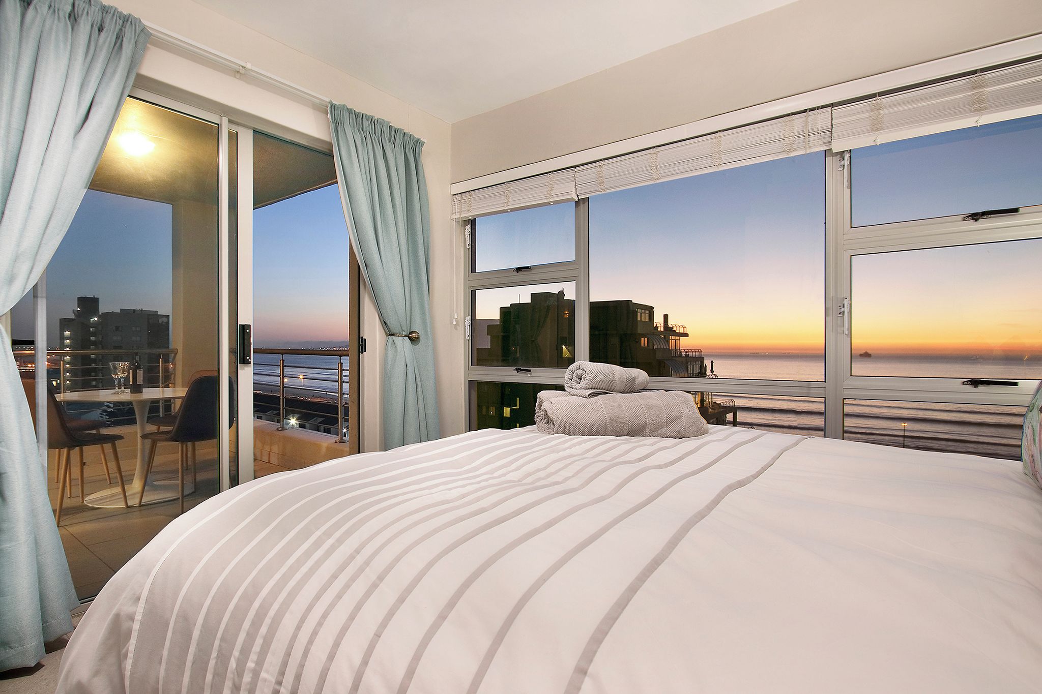 A hotel room with a large bed, a balcony with a view of the city, and a window with a sunset view.