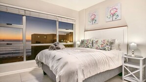 A bedroom with a large window, a bed with a white and grey bedspread, and two floral paintings on the wall. - Ocean View A603 (Cape Town)