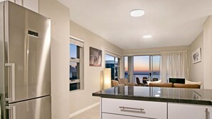 A modern kitchen with a stainless steel refrigerator, a granite countertop, and a view of the city at sunset through large windows. - Ocean View A603 (Cape Town)