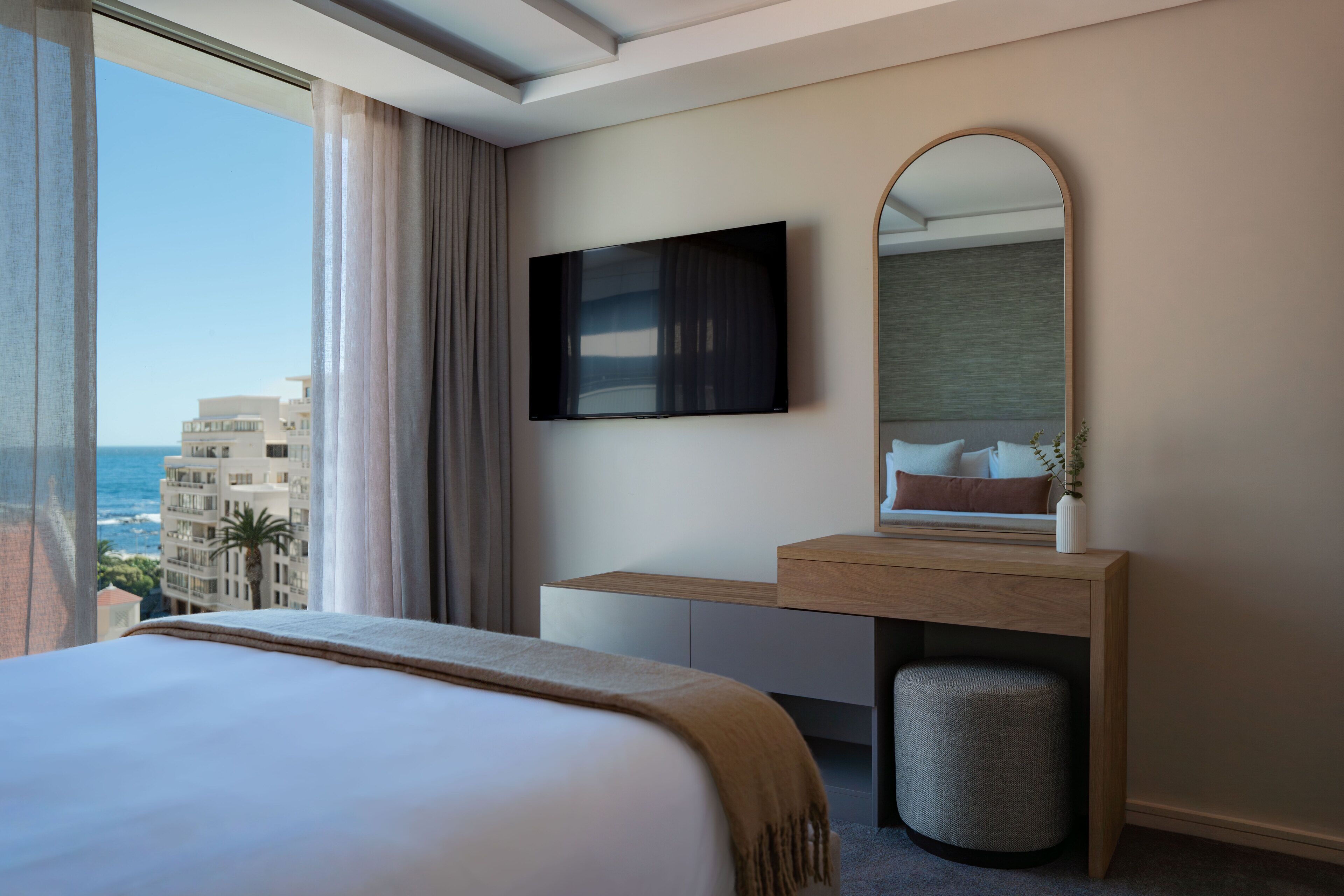 Ein Hotelzimmer mit einem Bett, einem an der Wand befestigten Fernseher, einem Holztisch, einem Spiegel und Blick auf das Meer und Gebäude.
