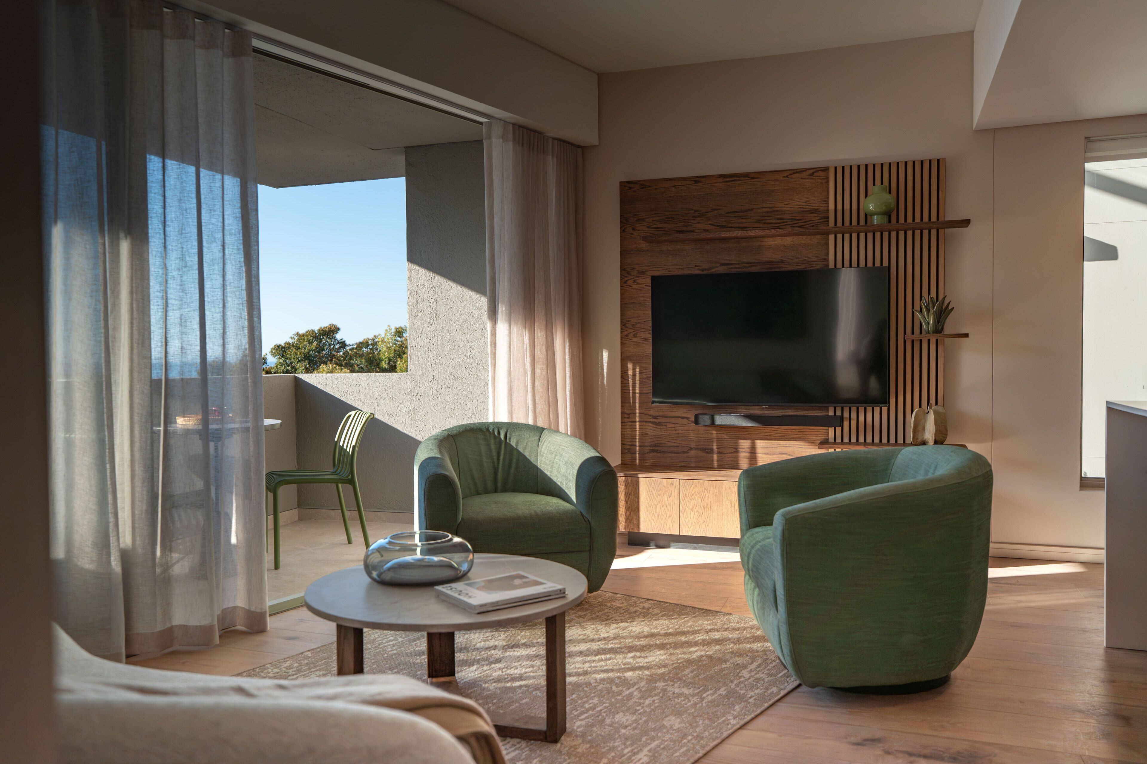A modern living room with a flat-screen TV, two green armchairs, a coffee table, and a sliding glass door leading to an outdoor area.