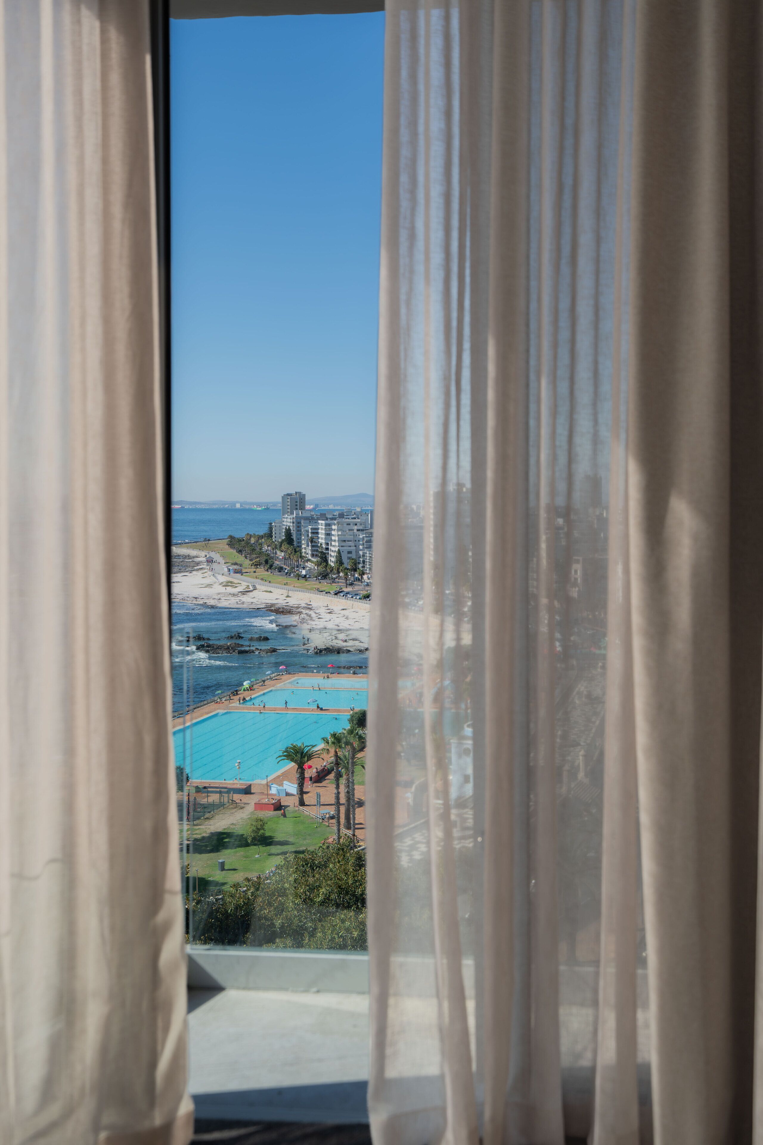 Vista a través de una ventana con cortinas translúcidas, que muestra una playa, el océano y un conjunto de edificios.