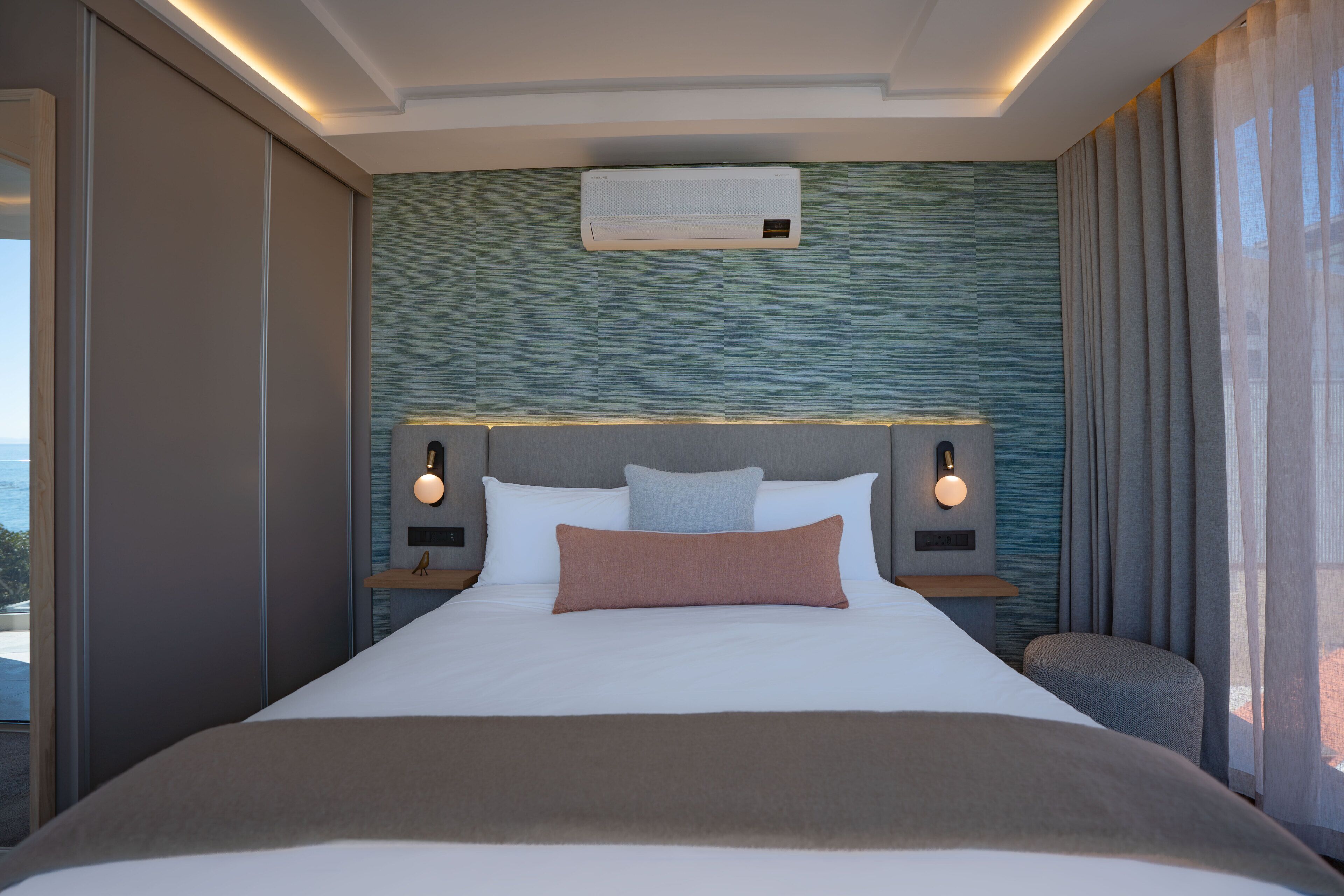A hotel room with a bed, an air conditioner, a nightstand with a lamp, and a view of the outdoors.