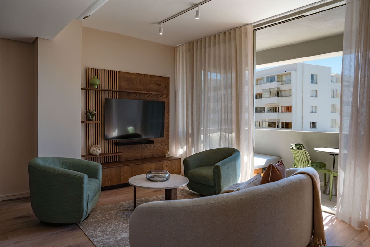 Un salon moderne avec un téléviseur à écran plat, deux fauteuils verts, un canapé, une petite table ronde et une vue sur un immeuble à travers des rideaux transparents.