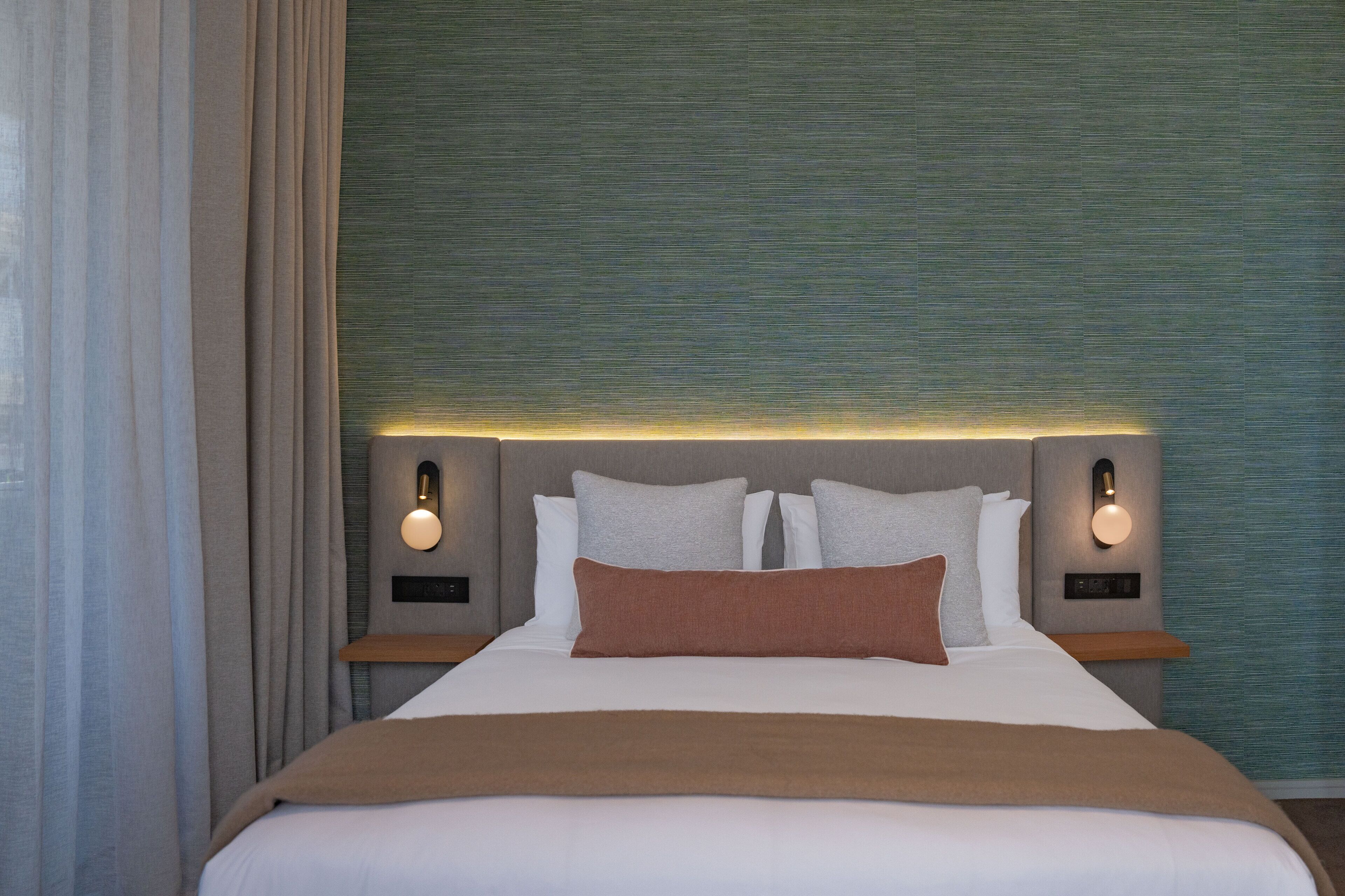 Una cama bien hecha con una manta marrón, dos mesitas de noche con lámparas de pared y una pared verde con textura.