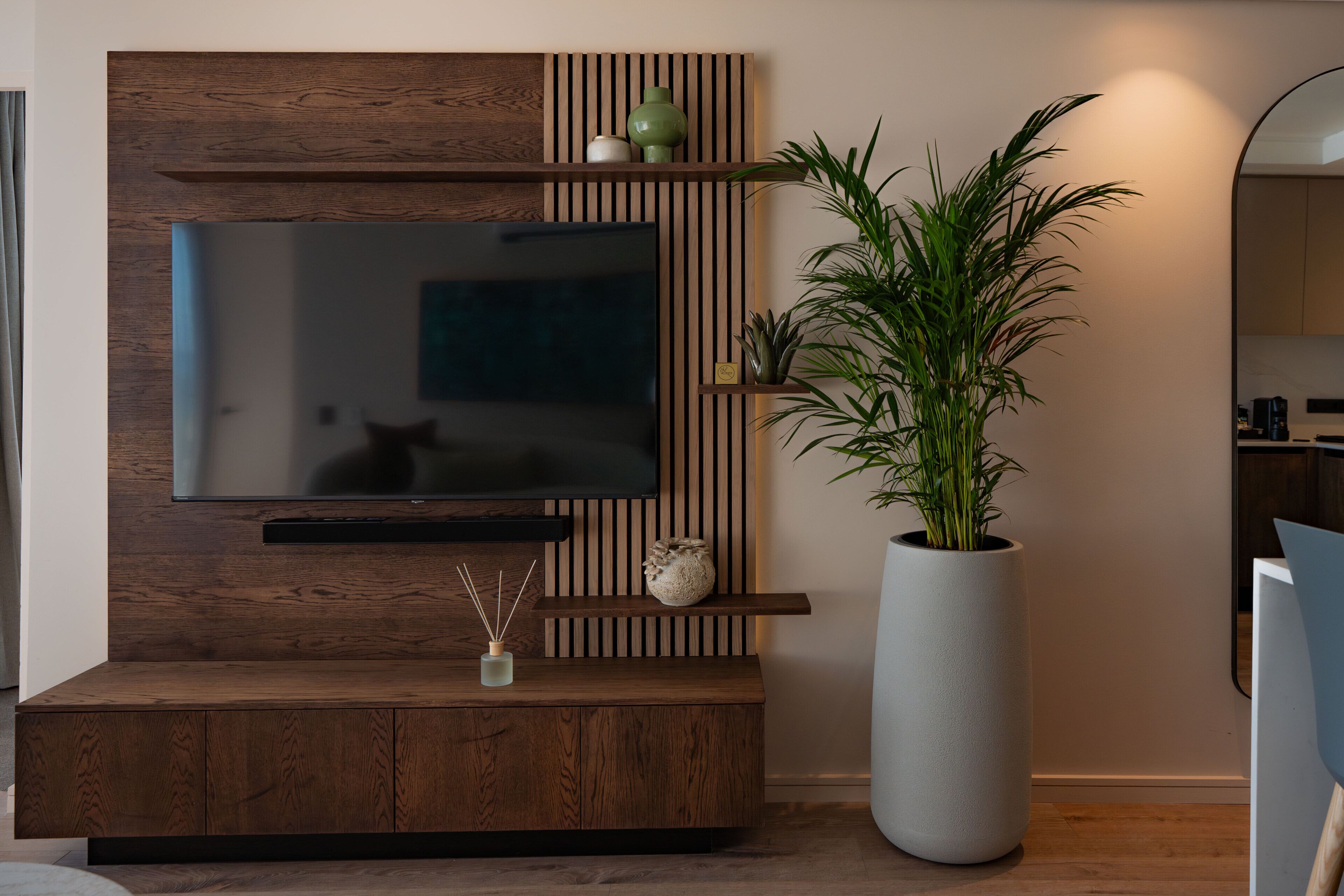 A modern living room with a flat-screen TV mounted on a wooden wall unit, a large potted plant, and a minimalist design.
