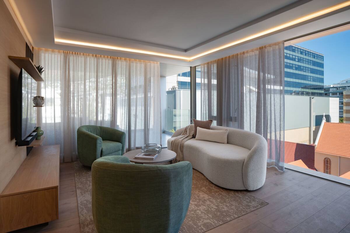 Un salon moderne avec un canapé, un fauteuil et une table basse, de grandes fenêtres avec des rideaux et une vue sur un immeuble.