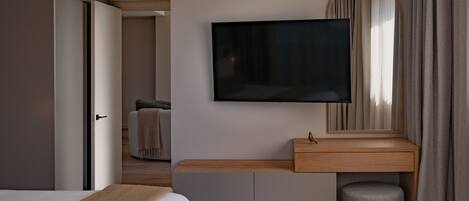 A modern hotel room with a bed, a TV mounted on the wall, a wooden bedside table, a mirror, and a small ottoman.