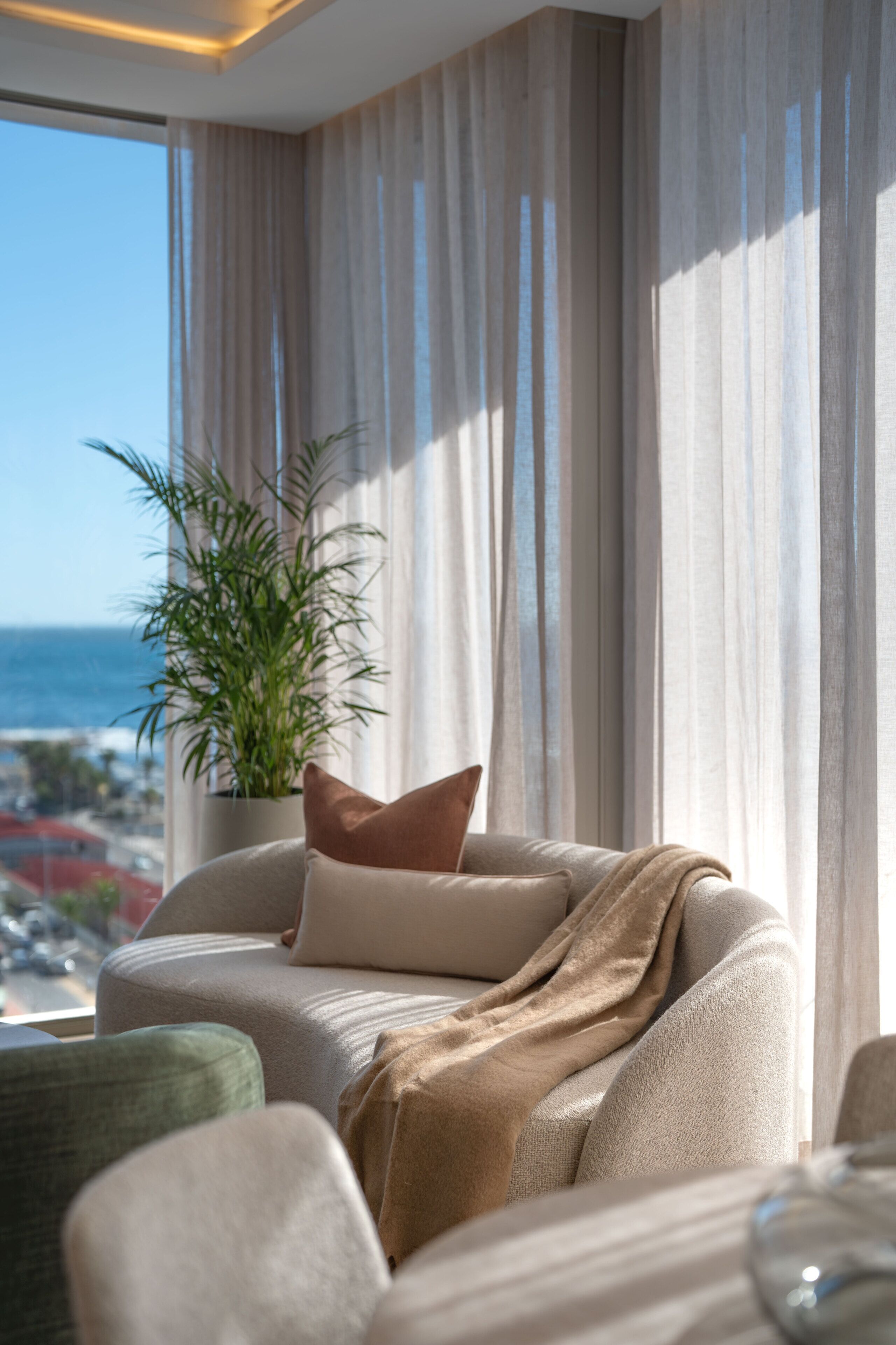A modern living room with a view of the ocean, a potted plant, and a sofa with a throw blanket.