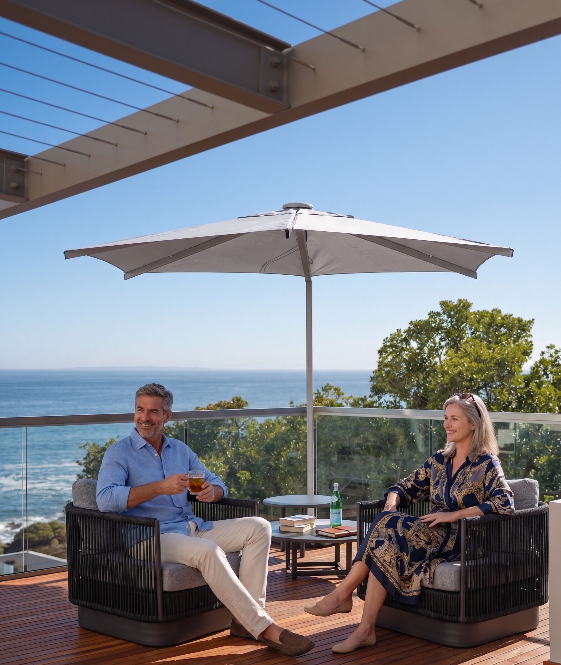 Deux personnes assises sur des meubles de jardin, avec vue sur l’océan, une table avec des livres et une bouteille, et un grand parasol.