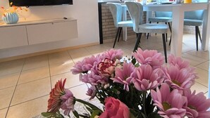 A modern living room with a flat-screen TV mounted on the wall, a dining area with blue chairs, and a large window offering a view of greenery.