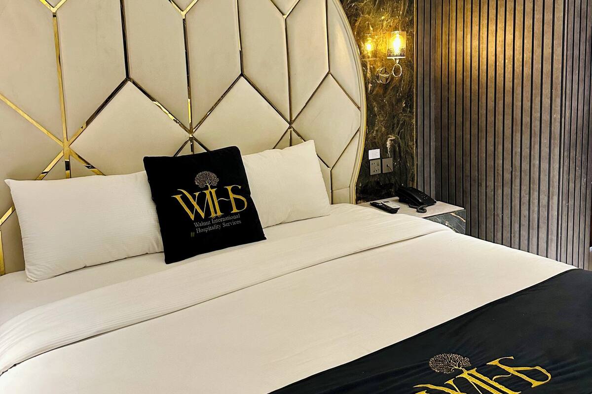 A hotel room with a large bed, a headboard with a geometric pattern, a black throw with gold lettering, and a wall-mounted lamp.