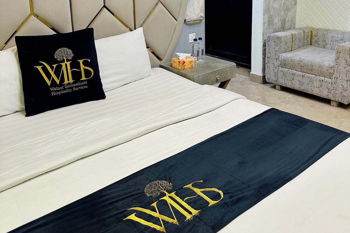 A hotel room with a large bed, a headboard with a geometric pattern, a black and gold pillow, a nightstand, a sofa, and a patterned wall.