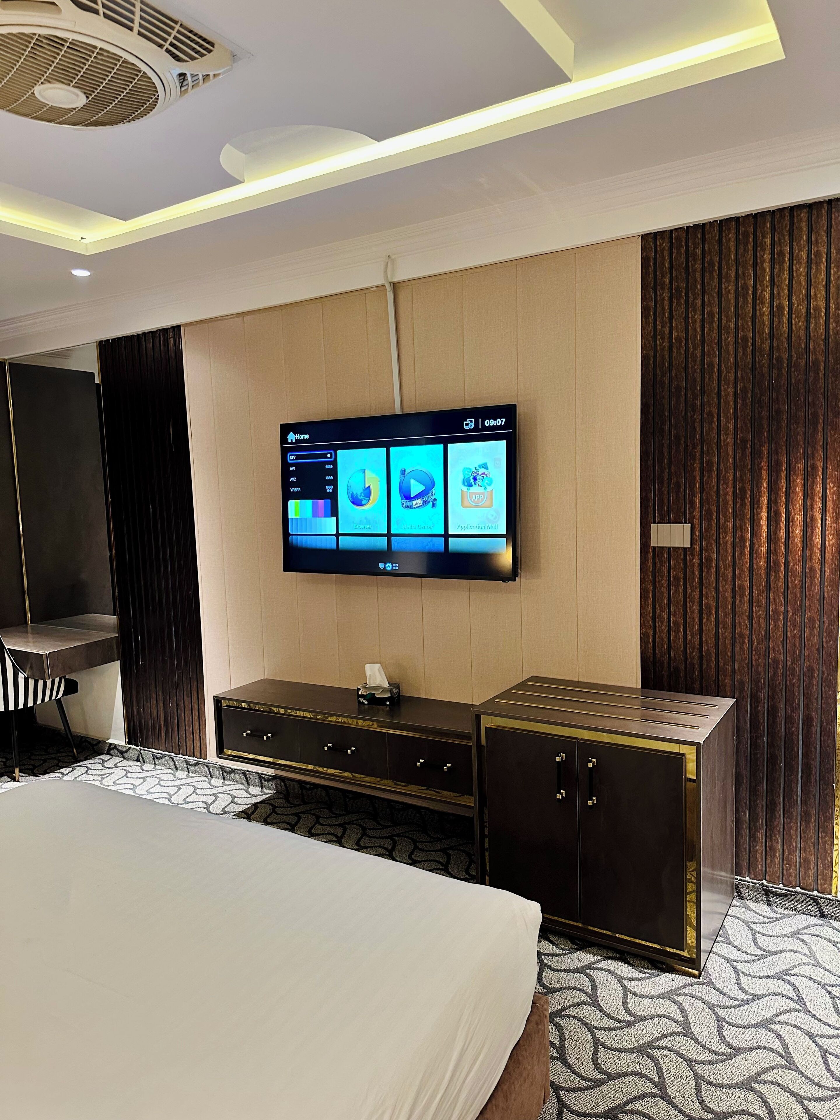 Une chambre d’hôtel moderne dotée d’un grand téléviseur à écran plat fixé au mur, d’un lit recouvert d’une courtepointe blanche, d’une commode en bois foncé et d’un tapis à motifs.