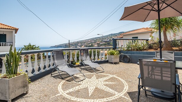 A rooftop terrace with a view of the sea, two lounge chairs, a table, and a large umbrella.