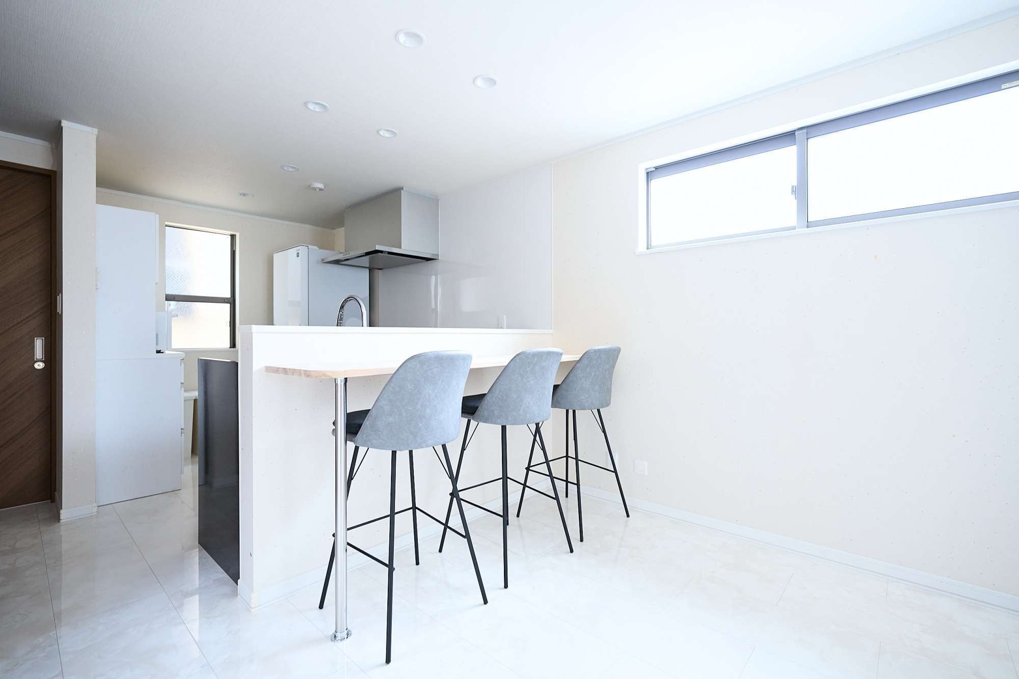 Une cuisine moderne avec un plan de travail blanc, quatre tabourets de bar gris et une fenêtre laissant entrer la lumière naturelle.