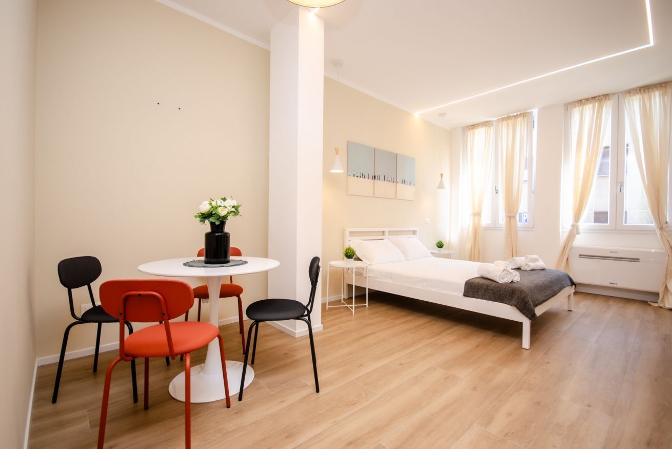Une chambre d’hôtel moderne, équipée d’un lit, d’une petite table avec des chaises, d’un vase de fleurs et d’une fenêtre avec des rideaux.