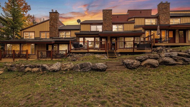 Una casa de dos pisos con una chimenea de piedra, un porche con mobiliario exterior y un muro de contención de piedra.