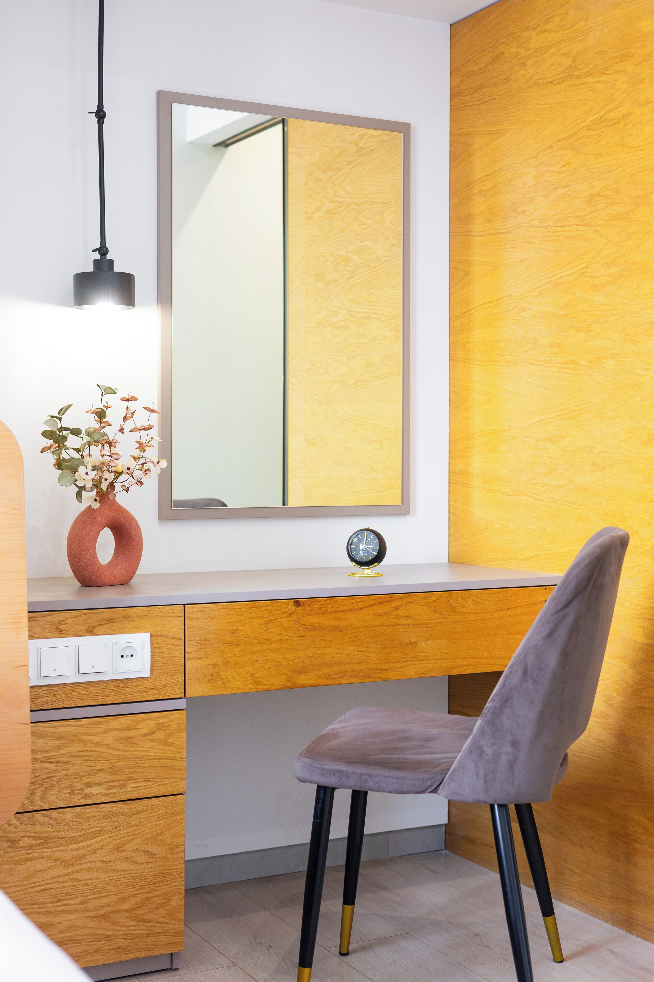 A modern office space with a wooden desk, a grey chair, a mirror, a vase with flowers, and a clock.