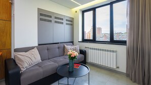 A modern living room with a grey sofa, a round black coffee table, and a vase with flowers. There is a window with a view of buildings and a radiator on the wall. - Apartment by Luxe Group KO333 (Yerevan)