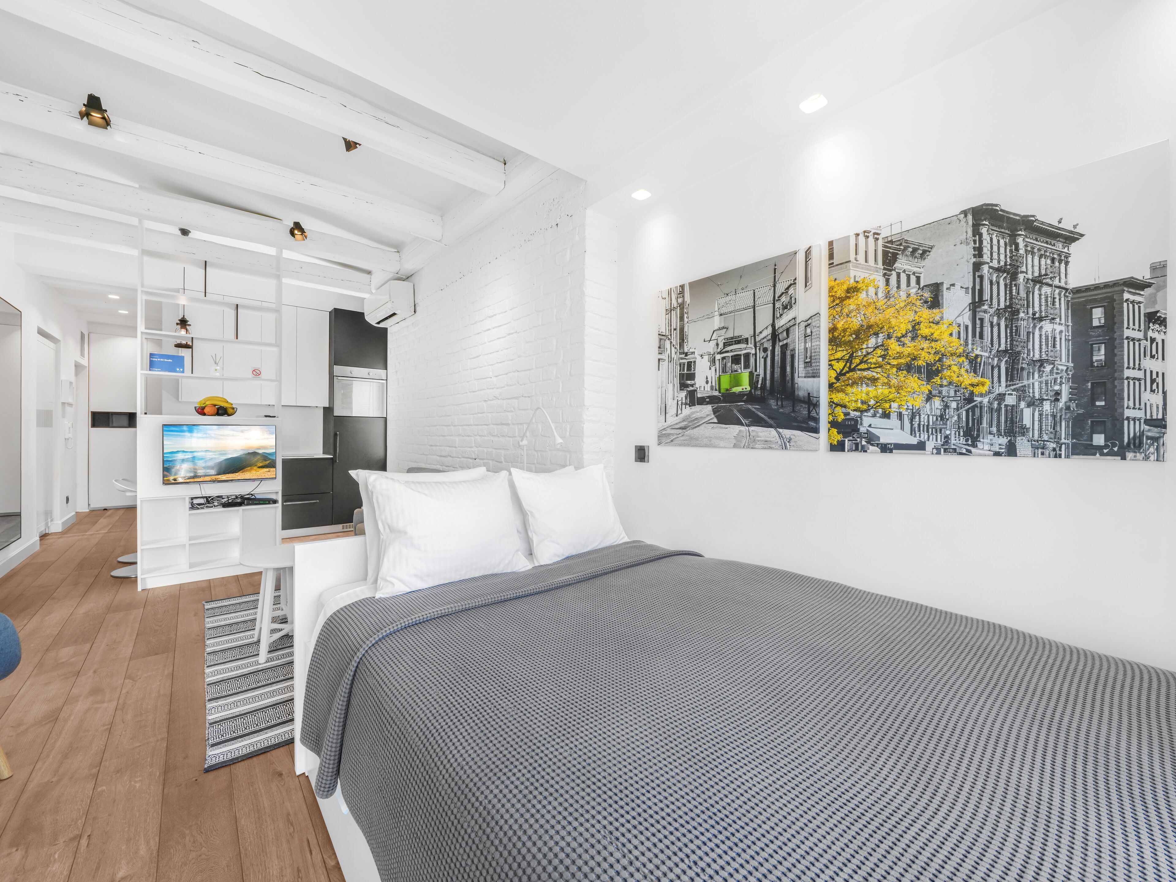 Ett modernt hotellrum med en säng, en tv, en vit tegelvägg och en tavla som föreställer en stadsbild.