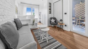 Ein modernes Wohnzimmer mit einem grauen Sofa, einem kleinen runden Tisch und einem grauen Sessel mit einem Sternenkissen. Ein Fenster mit weißen Vorhängen, eine weiße Ziegelwand und ein Blick auf einen Balkon mit Holzmöbeln.